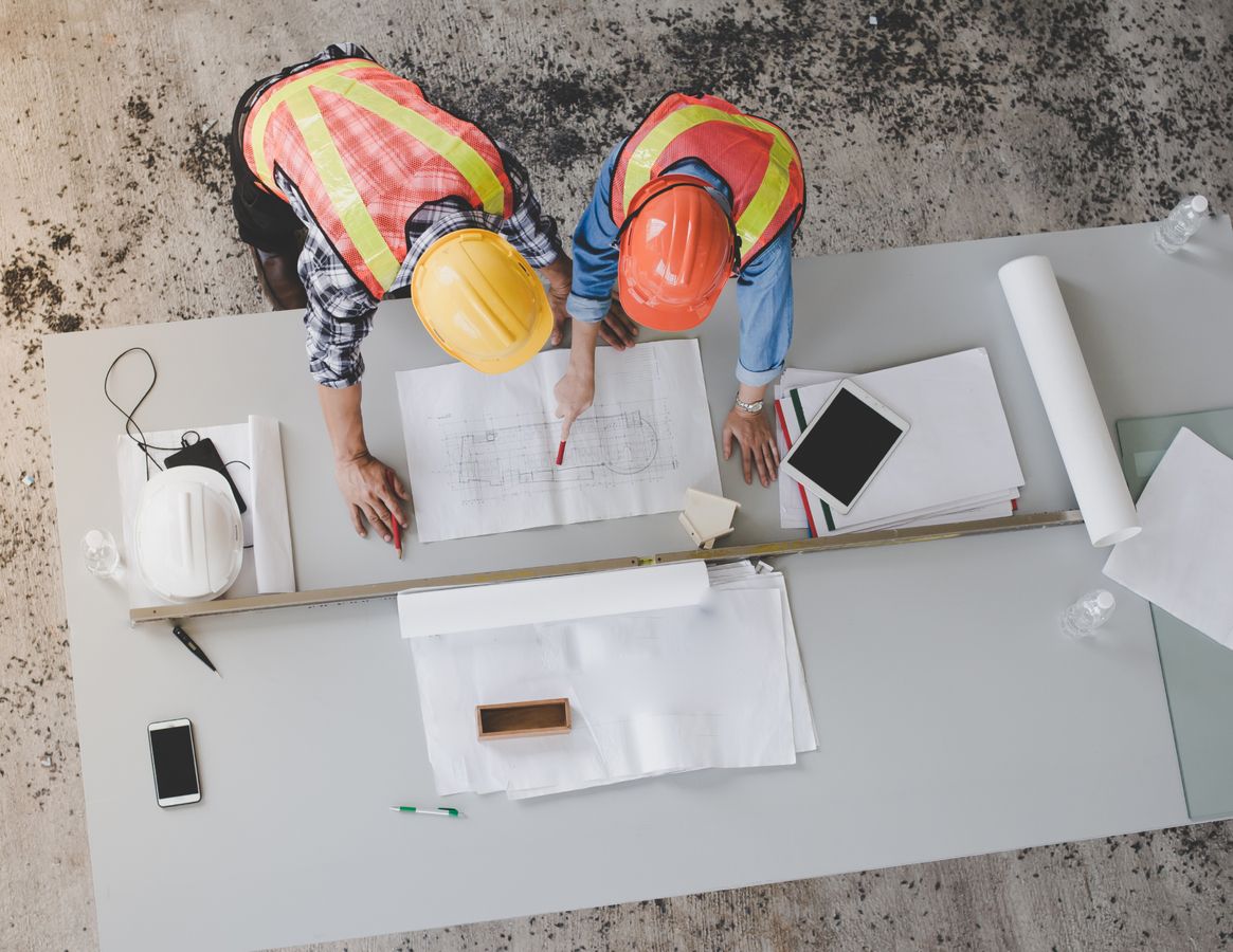 Zwei Personen in Schutzhelmen und Sicherheitswesten arbeiten an einem Tisch.